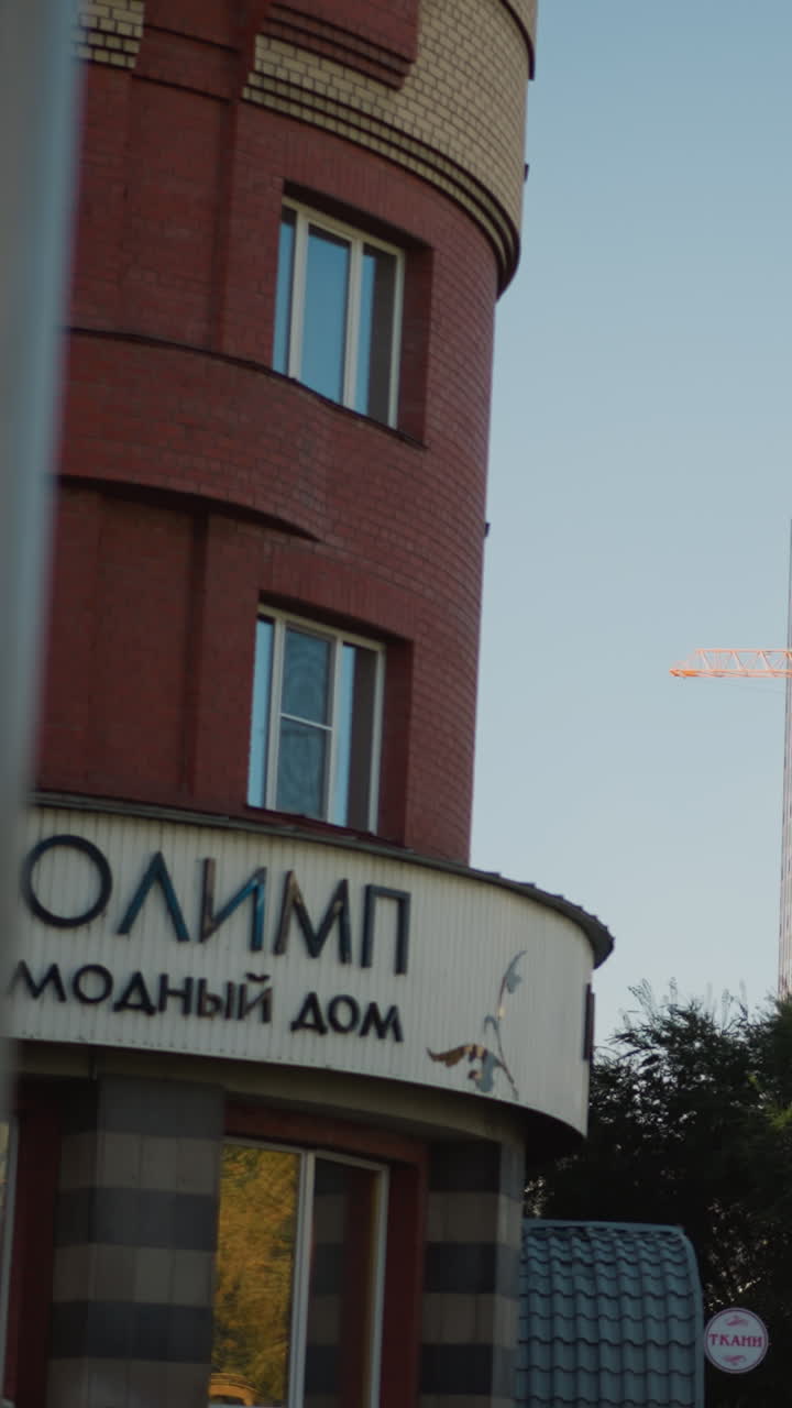 sunlit urban high rise and storefront. Warm late afternoon light illuminates apartment tower with construction crane crossing clear blue sky. Rounded red brick boutique facade displays Cyrillic