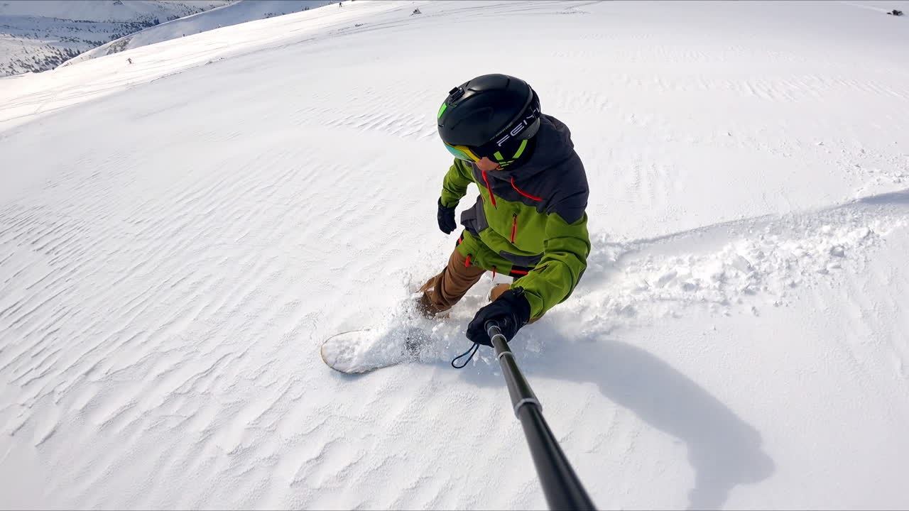 Man wearing sport suit and protective glasses rides down the hill on his snowboard. Snowboarder taking selfie video.
