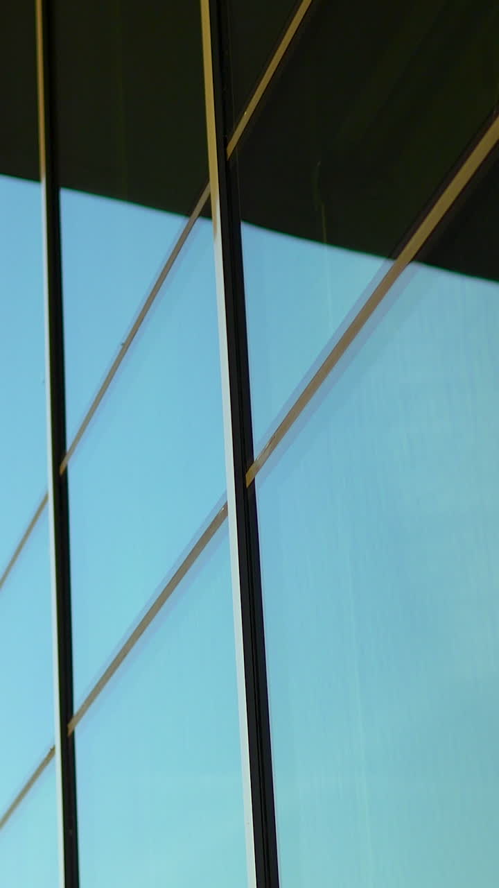 Una prospettiva verticale minimalista dell'esterno di un edificio per uffici contemporaneo, caratterizzato da pannelli in vetro geometrici e riflessi di cielo azzurro limpido. Ideale per contenuti aziendali e architettonici
