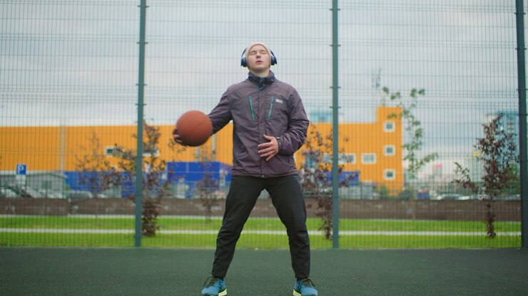 Active white man dribbling basketball outdoors, full body stance, tossing ball between hands, headphones on, focused gaze, fenced court with colorful industrial backdrop, amateur athlete performing