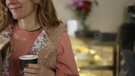 cajero masculino en delantal dando una bolsa de papel con pan a la mujer sonriente y ella se va sosteniendo una taza de papel con café.