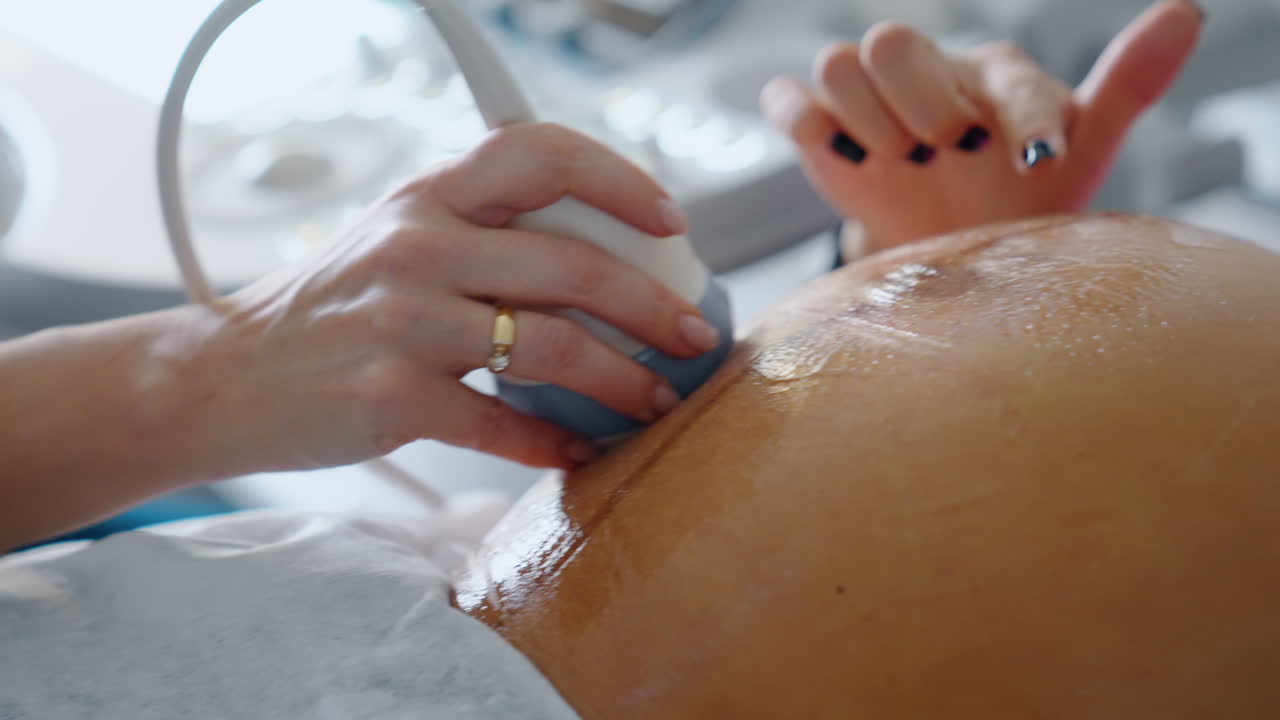 Female hand of doctor holding ultrasonic device on pregnant belly. Abdomen is covered with gel. Close up.