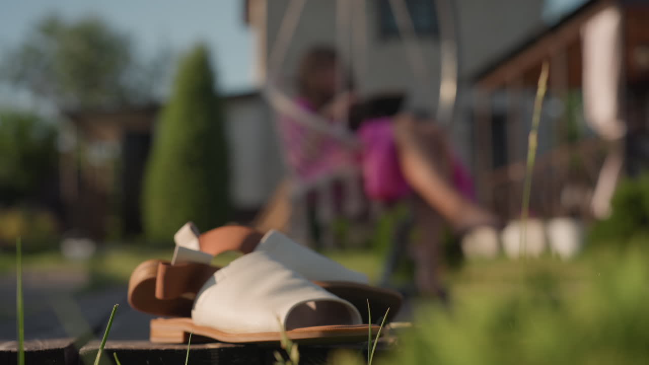 Woman Unwinding In Sunny Garden, Relaxed Lady Enjoying Warm Summer Breeze Outdoors On Swing, Peaceful Summer Scene With Woman Sitting On Swing Surrounded By Bright Sunlight And Sandals