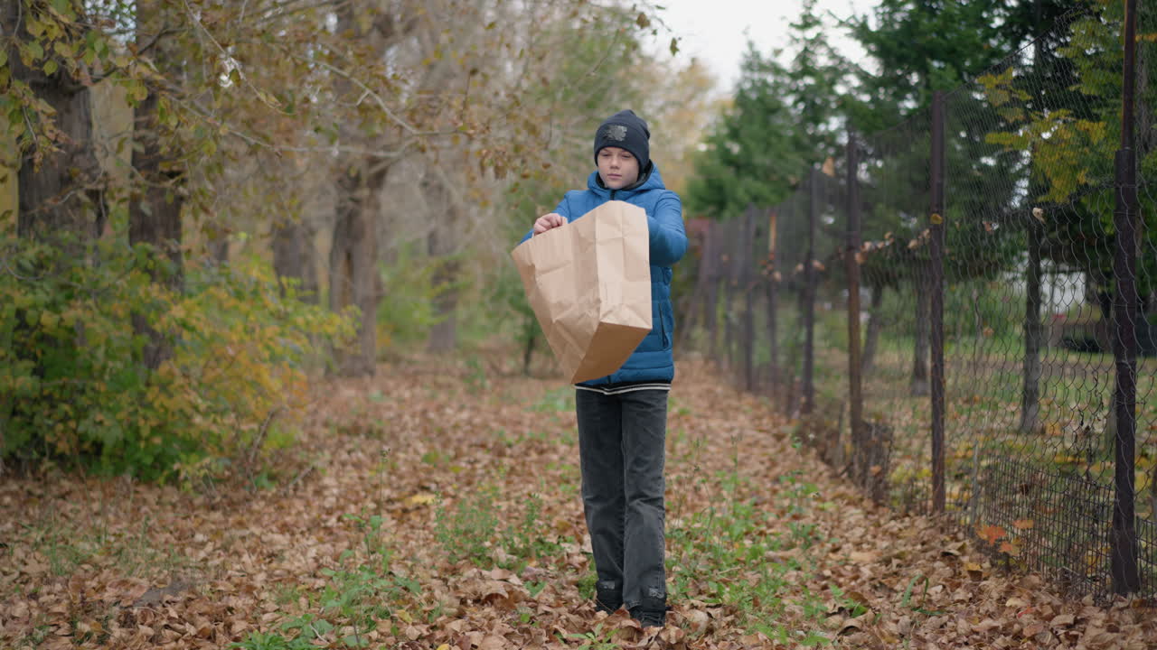 어린 아이가 건조한 잎을 즐겁게 고, 바게 종이 봉투를 거꾸로 돌려 땅에 건조 한 잎을 부어, 활기찬 잎자루와 시골 울타리 배경을 가지고 있습니다.