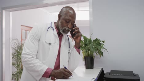 African-american-male-doctor-wearing-lab-coat-and-stethoscope,-talking-on-smartphone,-slow-motion