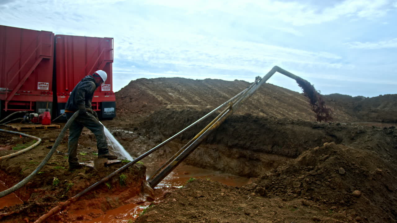 Man in white helmet and dirty clothes holding a hose with pouring water. Dirt is coming from a pipe into the special pool at oil or gas producing site.