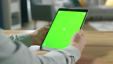 hombre usando gestos de la mano en pantalla de maqueta verde computadora tableta digital en modo retrato mientras está sentado en su escritorio. en el fondo acogedor salón.