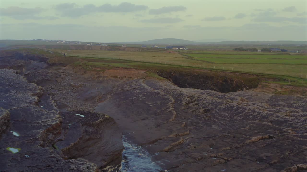 Sunset view over the Bridges of Ross, tranquil water reflecting warm light on the serene coastal scene as drone descends stratified rock sheets leading to grassu hills and arch