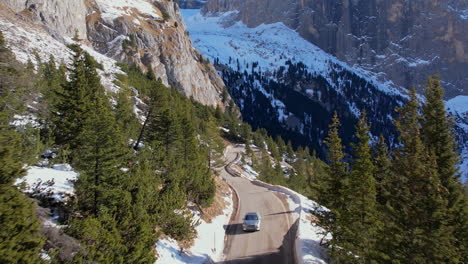 Una-Cautivadora-Toma-Aérea-De-Una-Carretera-De-Montaña-Que-Serpentea-A-Través-De-Un-Bosque-Nevado-En-Las-Dolomitas,-Con-Imponentes-Acantilados-Al-Fondo.