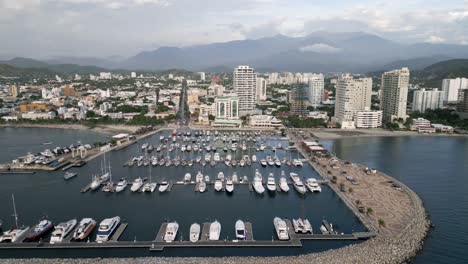 Flying-Above-Santa-Marta-City-Marina,-Colombia,-Aerial-View-of-Sailboats,-Breakwater-and-Cityscape-Skyline