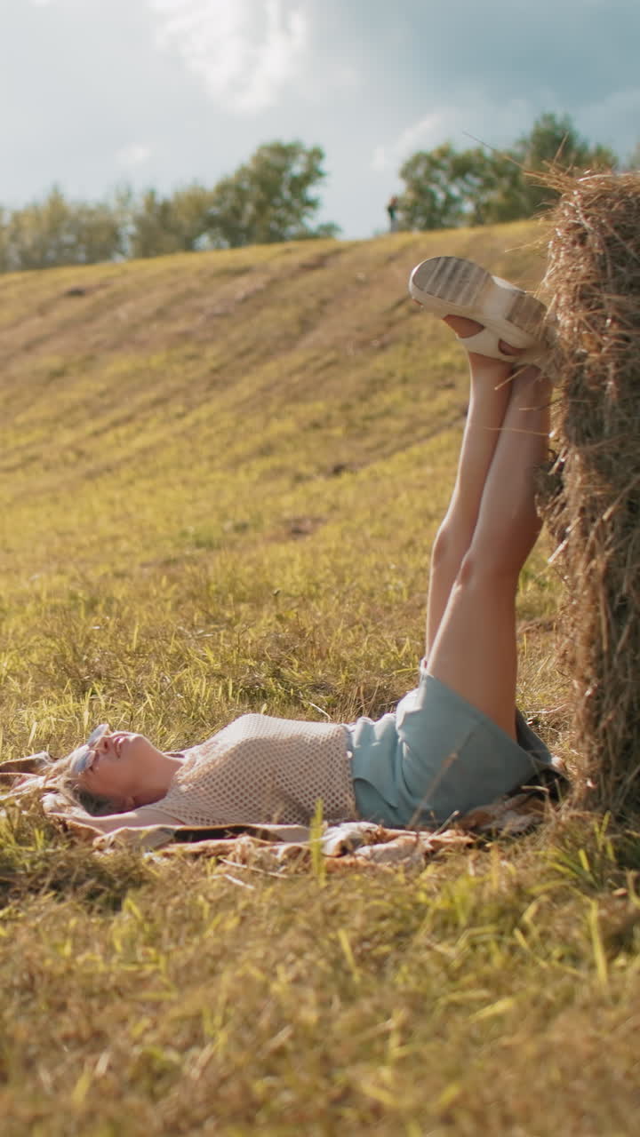 Lady taking sister photos outdoors as she relaxes on mat, placing legs on hay in vast farmland, warm sunlight, countryside scenery, casual photography, and rustic setting with hat resting on hay bale