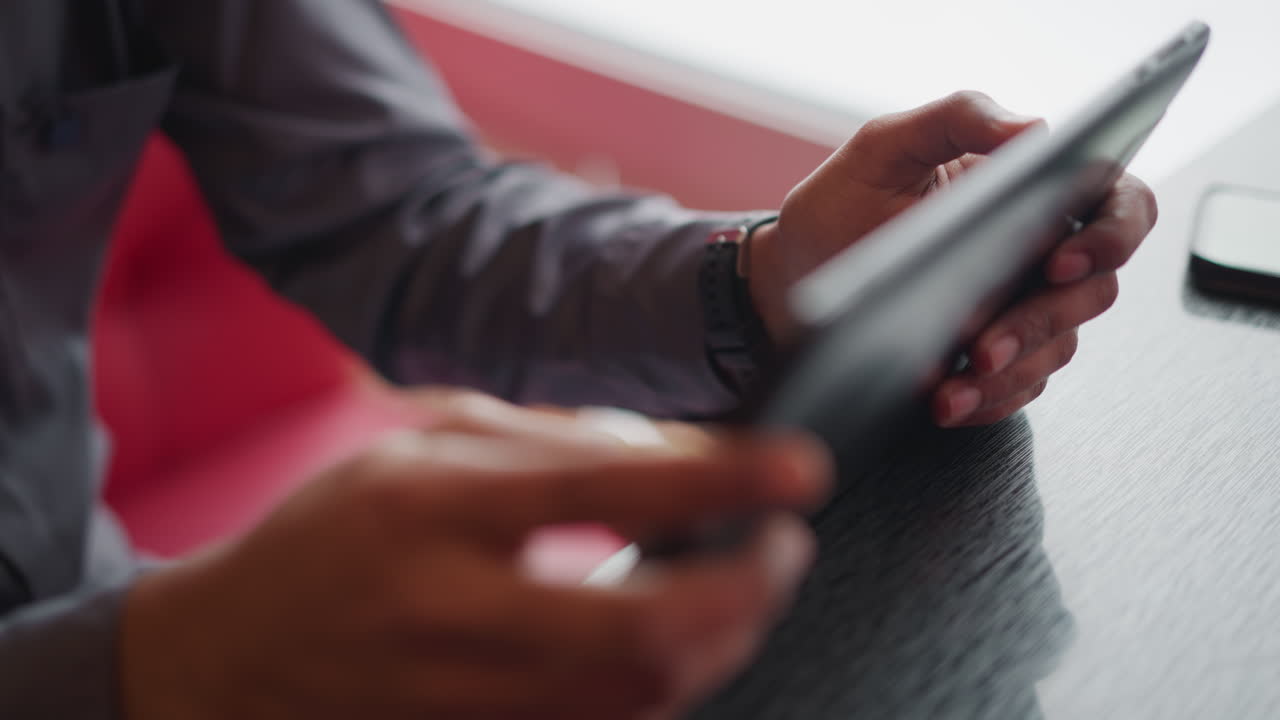 Immigrant using tablet indoors near window, hands holding and interacting with screen in close-up view, dressed in casual shirt, absorbed in digital task or browsing activity in relaxed modern setting