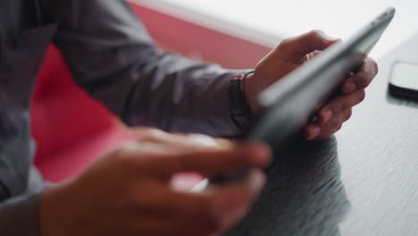 immigrant using tablet indoors near window, hands holding and interacting with screen in close-up view, dressed in casual shirt, absorbed in digital task or browsing activity in relaxed modern setting