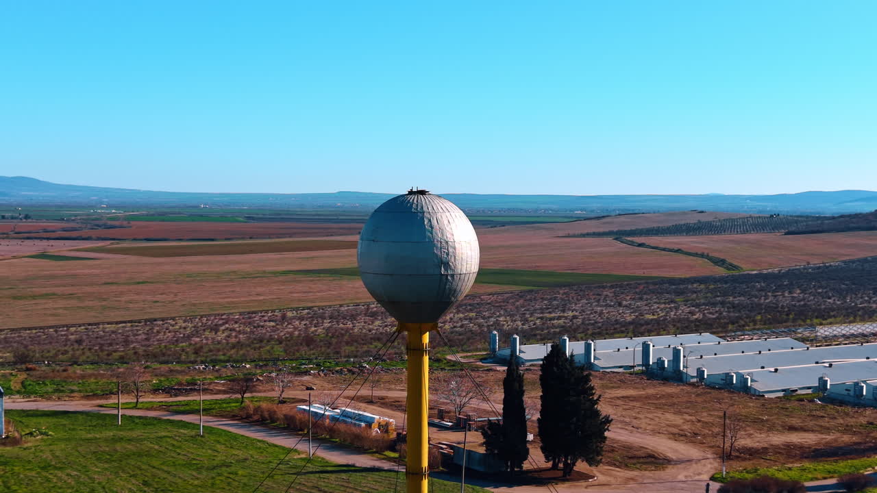 Torre d'acqua sferica presso una struttura agricola in zona rurale. Serbatoio d'acqua sferico in metallo su pilastro giallo che domina fienili agricoli e vasti campi sotto un cielo blu