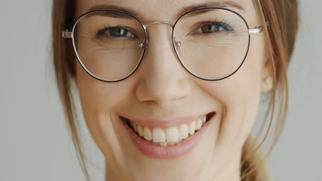 close up of the woman's face in glasses with fair hair looking straight in the camera and smiling on the white wall background