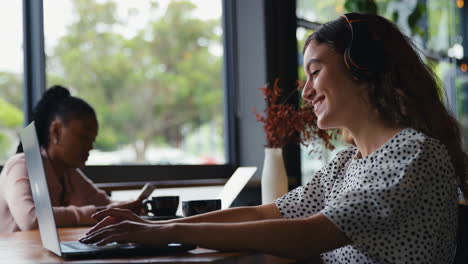 businesswoman wearing wireless headphones working on laptop in cafe with colleague in background