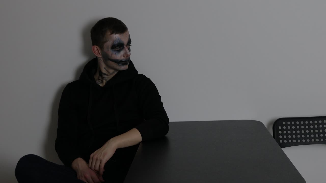 A man with dark clown makeup sitting alone and looking towards the wall while listening and being thoughtful
