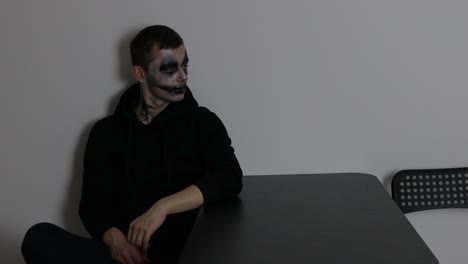 a man with dark clown makeup sitting alone and looking towards the wall while listening and being thoughtful