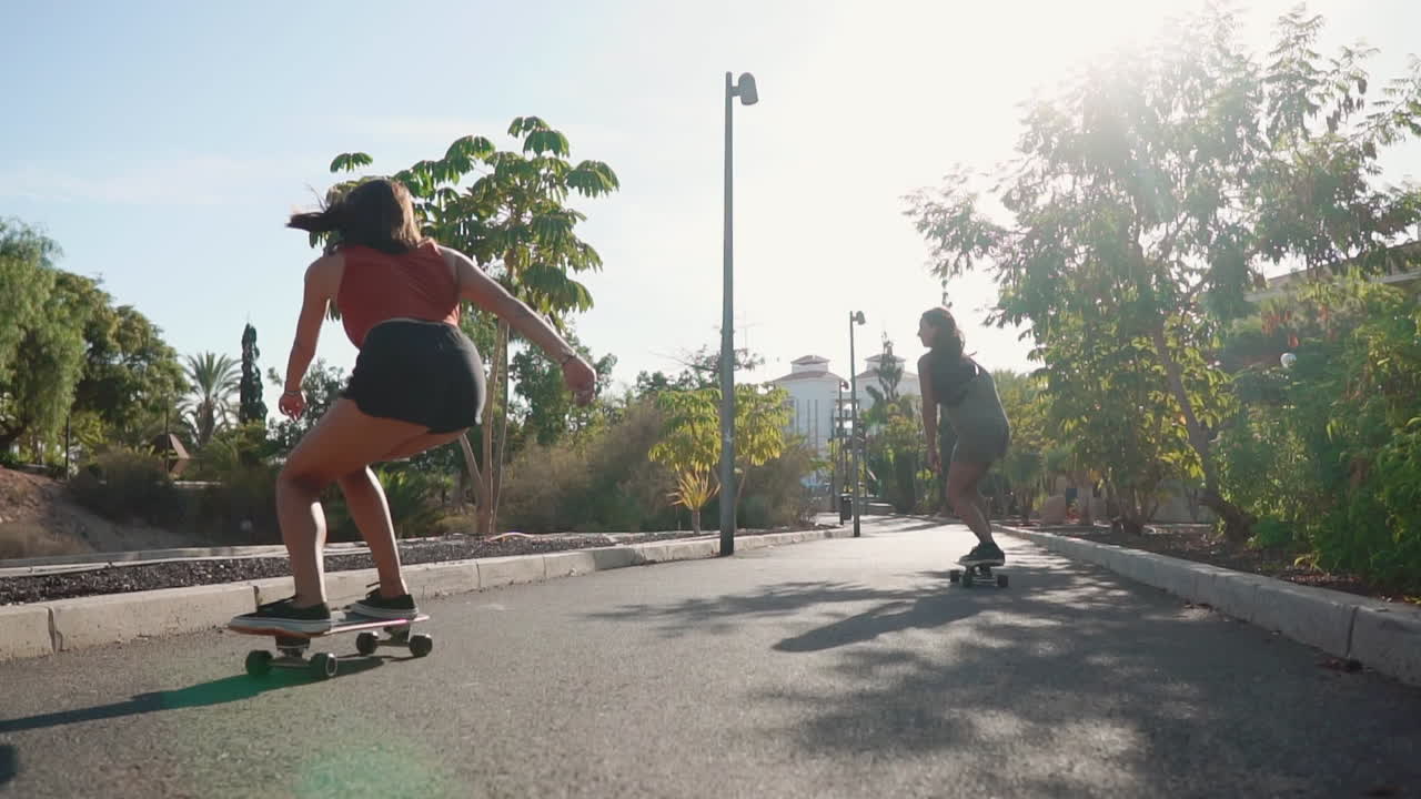 Amid palm trees, two young Hispanic women skate unhurriedly on island paths, their motions depicted in slow motion against the sunset's vibrant backdrop. This illustrates happiness and a commitment to health