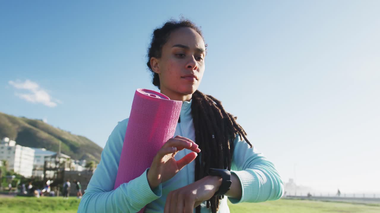 Mixed race fit woman holding pink yoga mat and checking her smartwatch outdoors
