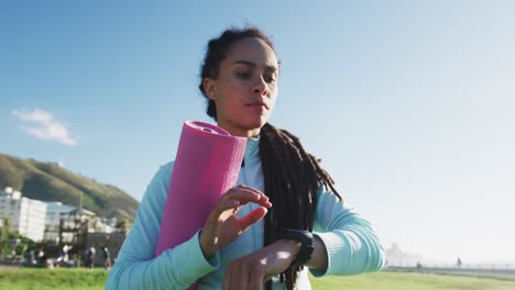 Mixed-race-fit-woman-holding-pink-yoga-mat-and-checking-her-smartwatch-outdoors