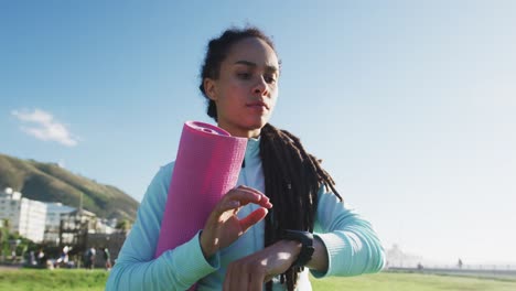 mixed race fit woman holding pink yoga mat and checking her smartwatch outdoors