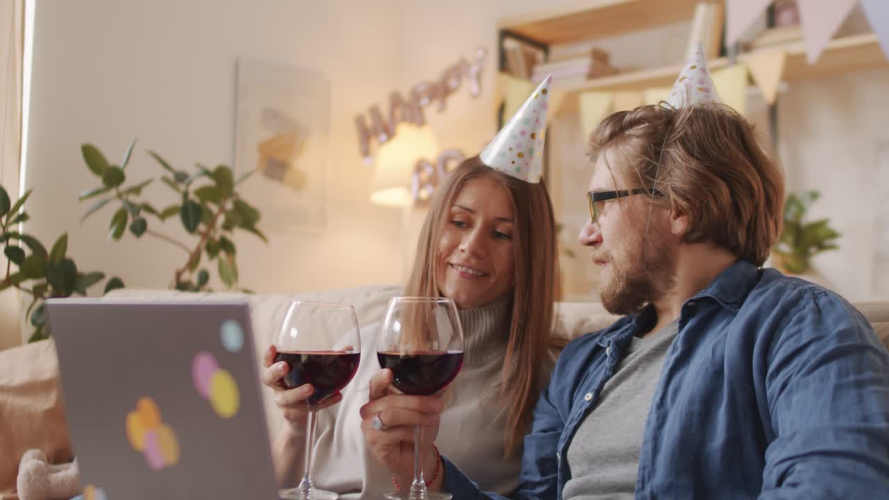 casal comemorando aniversário online usando chapéus de aniversário e brindando com taças de vinho tinto
