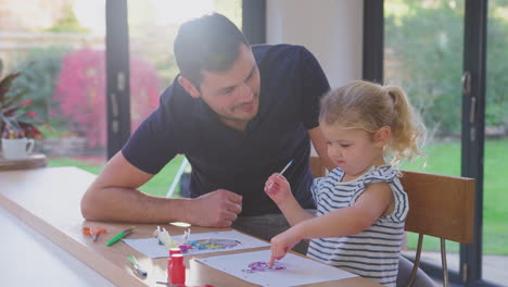 Father-and-young-daughter-having-fun-at-home-sitting-at-table-and-painting-decoration-together---shot-in-slow-motion