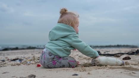 a blonde baby girl picking up a plastic bottle on the beach and throwing it at the camera