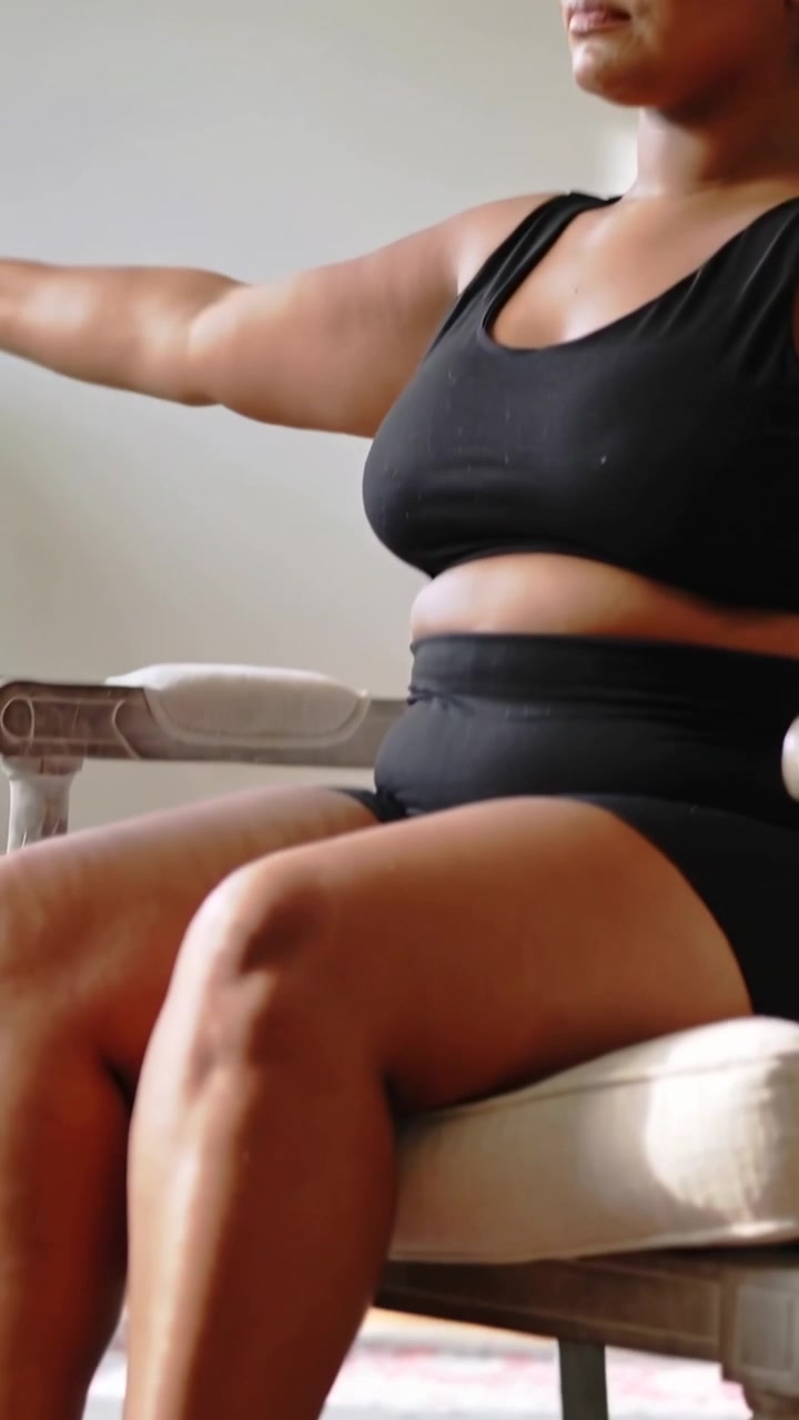 Video verticale: Donna che inspira muovendo le braccia, seduta a casa su un tappetino per sedia, in reggiseno e pantaloncini neri mentre si allunga