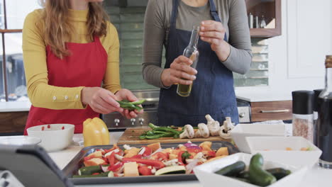 Happy-caucasian-lesbian-couple-preparing-food-and-using-tablet-in-sunny-kitchen