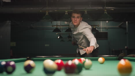 close-up of billiard balls on green pool table as player in white shirt and grey trousers leans over, gripping cue stick with intense focus. overhead lights cast dramatic shadows