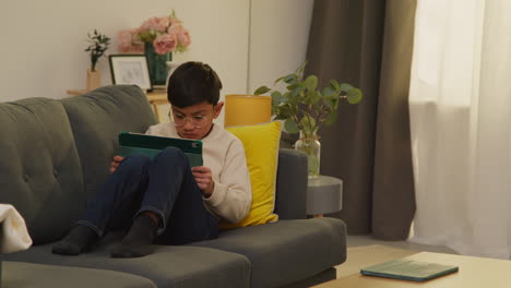 young boy sitting on sofa at home playing games or streaming onto digital tablet