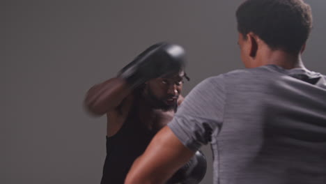 studio shot of male boxer sparring working out with trainer wearing punch mitts or gloves practising for fight 2