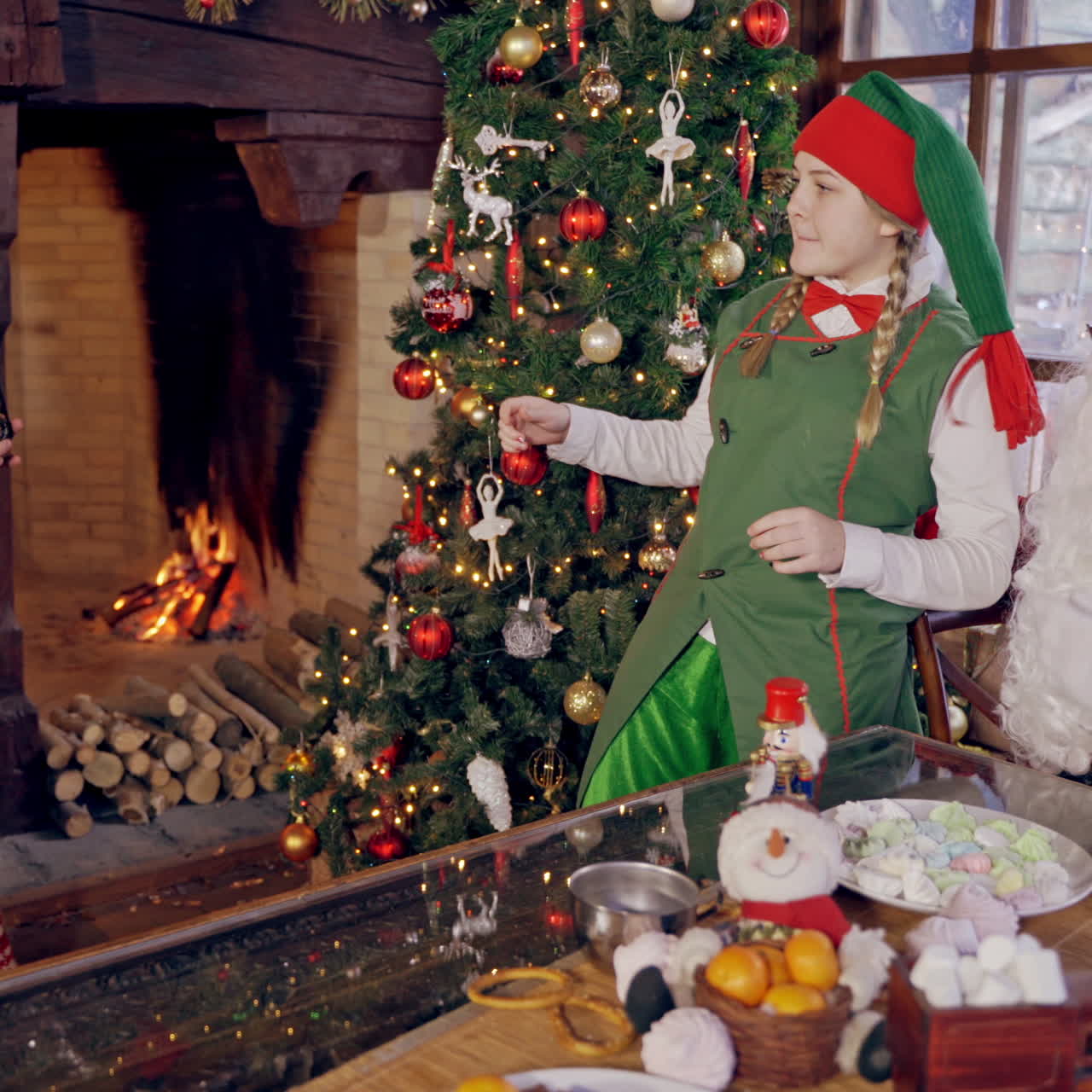 Santa with elf near the Christmas tree. Happy elf taking photo by retro camera on burning fireplace background. Happy xmas holidays.
