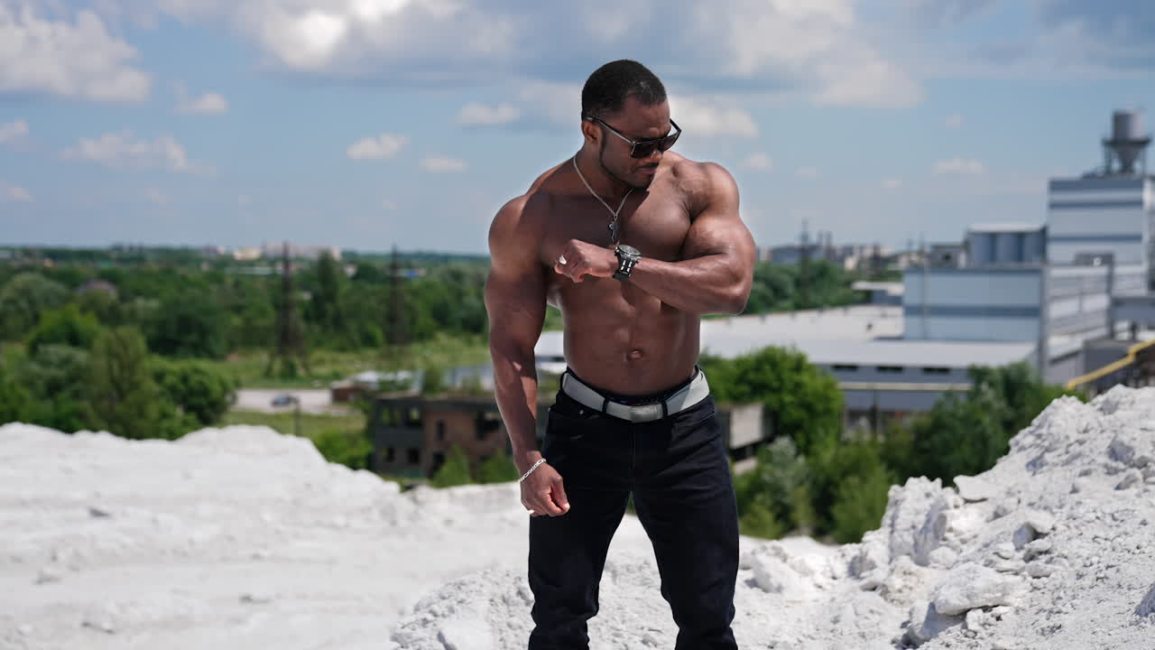 Male bodybuilder flexing muscles. African american sportsman standing shirtless showing muscles outdoors. Dark-skinned athlete in black glasses shows strong arms.