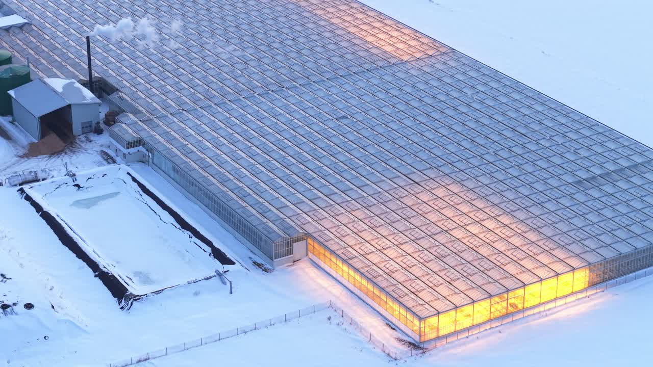 Nærbilde luftfoto tele av opplyst varmt glassdrivhustak i snødekt vintergårdslandskap luftfoto, med oppvarmet interiør
