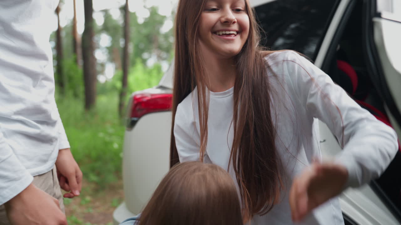 Beautiful girl carries younger sister down from car with bright smile surrounded by green forest, sunlight reflecting on white vehicle, capturing warmth of family bonding, and joyful outdoor travel