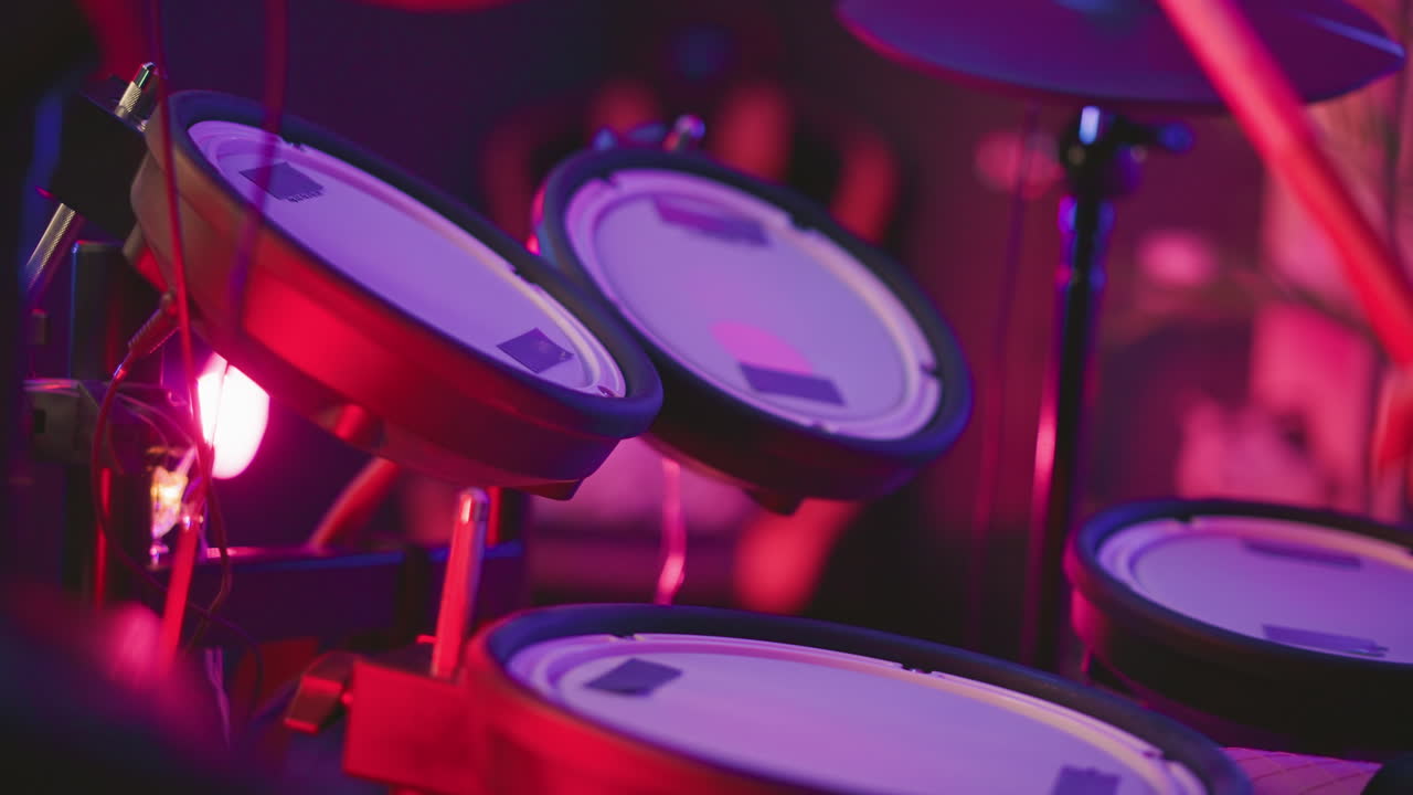 Close up of person playing electronic drum pads with sticks under vibrant red and purple lighting in indoor studio, mood dynamic and creative, blurred background hints neon glow and equipment