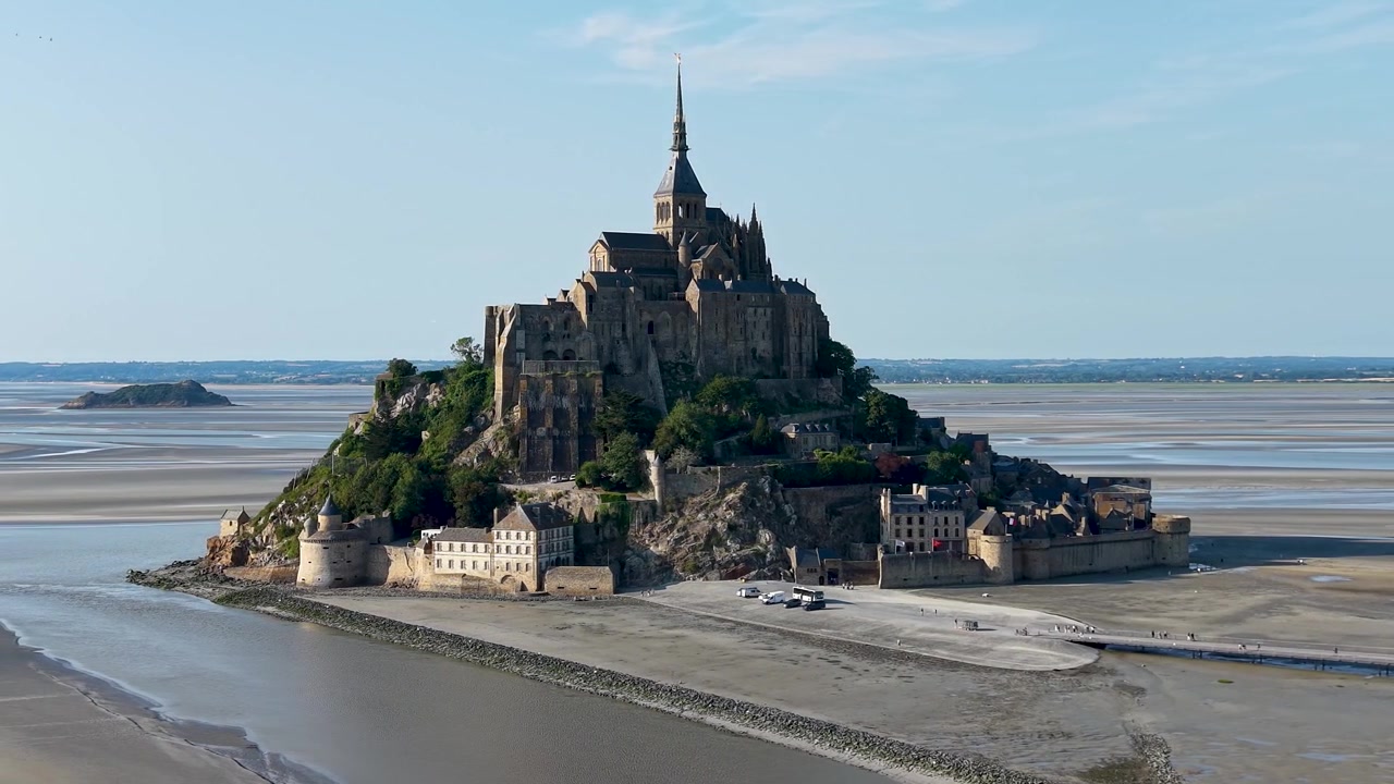 Vista aérea cinematográfica de drone em órbita lenta de Mont Saint-Michel, circulando graciosamente da direita para a esquerda ao redor da icônica abadia medieval, exibindo a ilha fortificada, as muralhas históricas e os bancos de areia