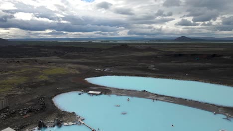 disfruta de la tranquilidad aérea sobre los baños naturales de mývatn, donde las piscinas calientes de color azul profundo ofrecen un sereno contraste con la extensión del lago mývatn en vívidos 4k