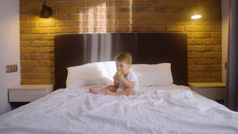 front view of a baby sitting on the bed in bedroom while bitting a green apple