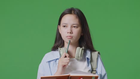 close up of asian teen girl student with a backpack taking note on notebook and thinking while standing in the green screen background studio