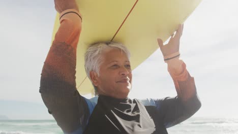 mujer hispana mayor feliz caminando por la playa con una tabla de surf sobre la cabeza