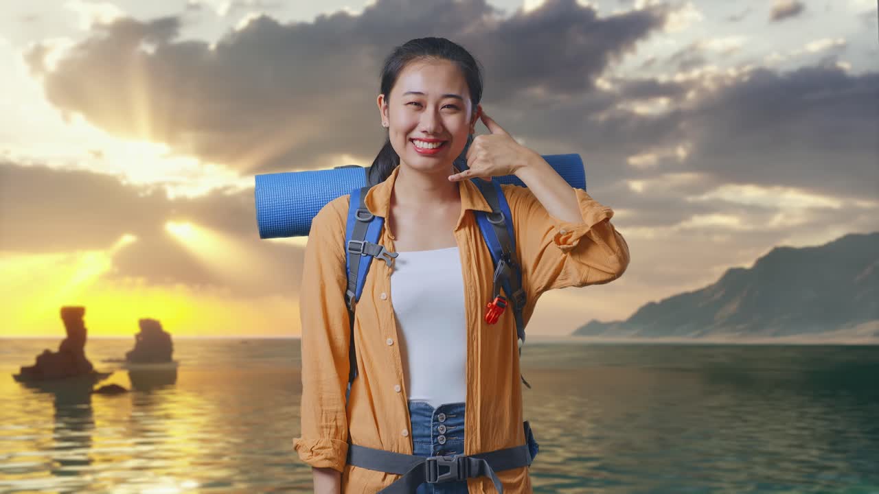 登山用バックパックを背負い、夕暮れ時のビーチに立って笑顔で電話をかけているジェスチャーをするアジア人女性ハイカー