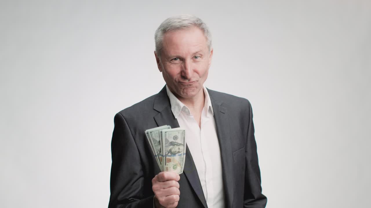 성공적인 은퇴와 부. 부유하고 자신감 넘치는, 슈트를 입은 노인이 카메라에 돈 한 어리를 보여주고 미소 짓고 있다.