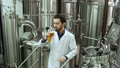 professional brewery worker in lab coat examining glass and trying freshly brewed beer at brewing factory