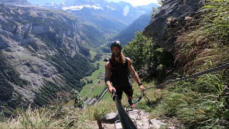 young, fit and athletic man is smiling to the camera while he slowly moves the camera to show the epic and beautiful landscape background behind him during his hike down via ferrata near murren.