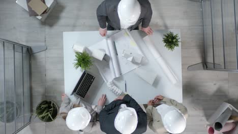 above view of two architects in safety helmets standing at table shaking hands and having talk concerning blueprint