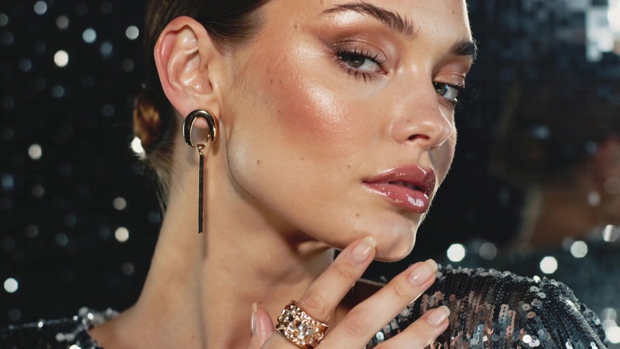 Modèle inclinant la tête, montrant une boucle d'oreille pendante, une bague, un haut à paillettes et un maquillage brillant, pendant que la caméra tourne en studio.